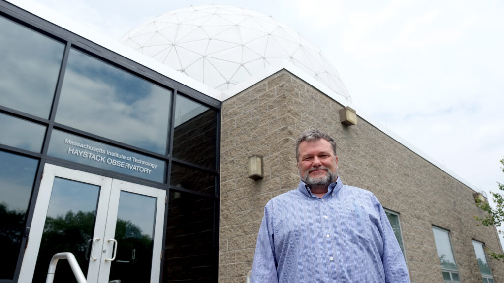 a man standing outside a large white radome next to a door labeled "MIT HAYSTACK OBSERVATORY"