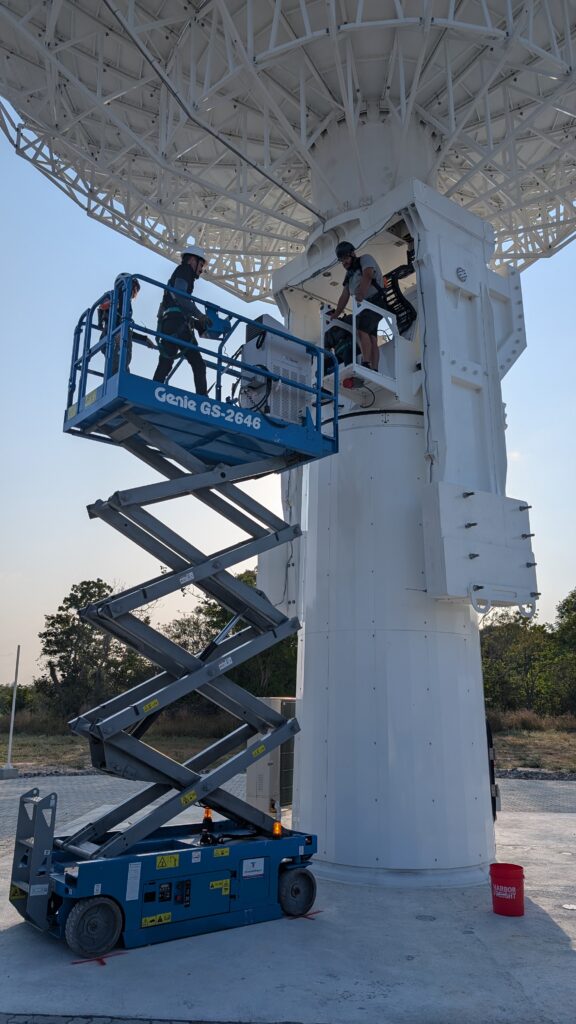 People on a scissor-style lift with a compressor going up to the middle of a large, white radio antenna