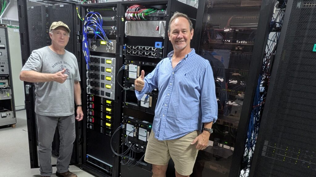 Two men each giving a thumbs-up next to racks of computer equipment