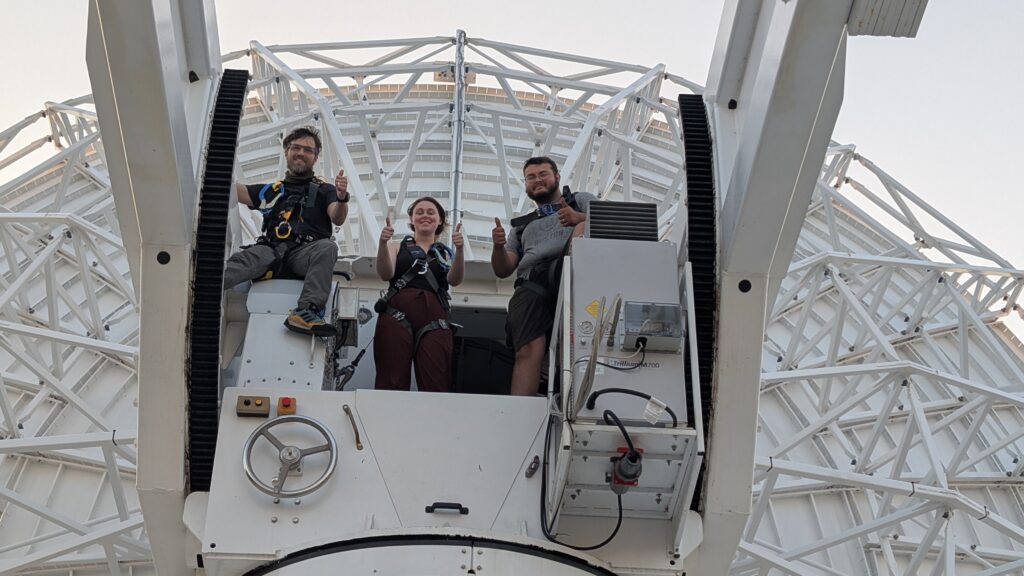 Three people each looking down and giving a thumbs-up from a platform outside a large, white radio antenna dish