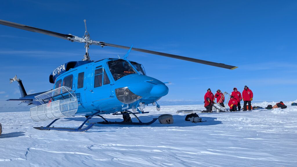 A blue helicopter resting on ice and snow in front of a group of people in red winter coats