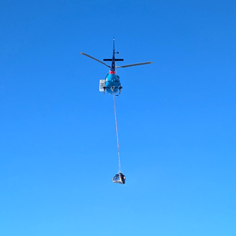 A helicopter carrying an item at the end of a long cable dangling underneath the helicopter
